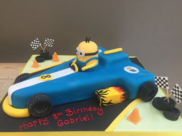 Birthday cake shaped like a blue race car with a Minion character, surrounded by racing elements on a gray background.