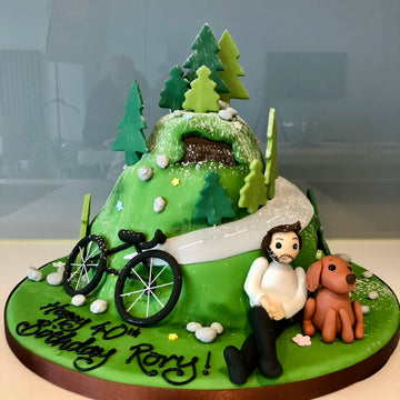 Birthday cake with a mountain, cyclist, and dog figure, decorated with trees and a bike.