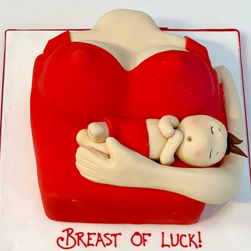 cake carved to the shape of a woman's chest with a baby held across her, labeled 'Breast of Luck', on a white background.
