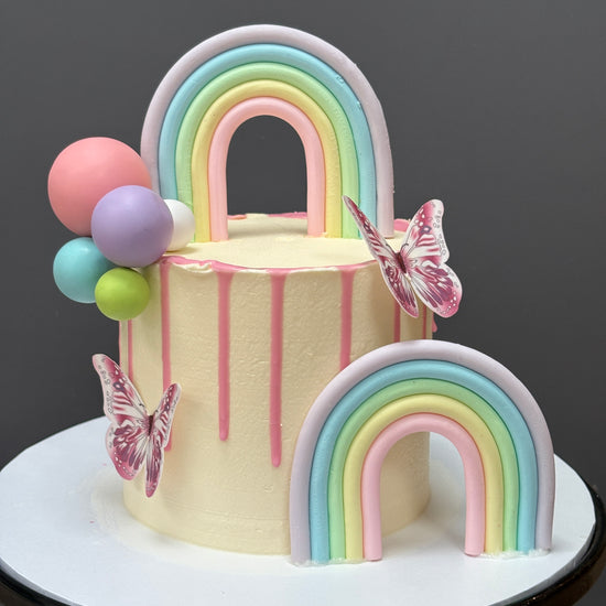 A pastel-colored frosted birthday cake on a white round stand, decorated with two fondant rainbows, pink butterfly embellishments, and pink, blue, purple, green, and pink balloon accents.