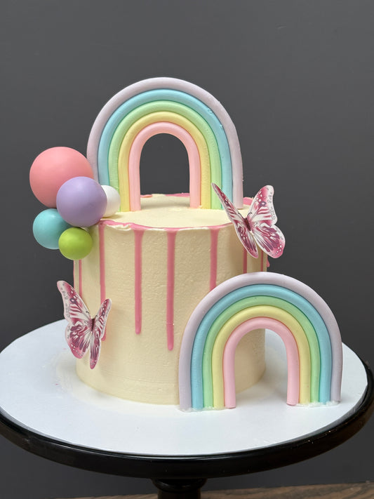 A pastel-colored frosted birthday cake on a white round stand, decorated with two fondant rainbows, pink butterfly embellishments, and pink, blue, purple, green, and pink balloon accents.