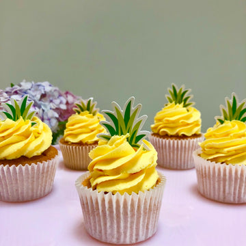 Cupcakes with yellow frosting and pineapple decorations on a light pink surface.