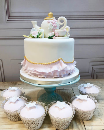 Decorative cake with teapot and flowers on a stand, surrounded by cupcakes.